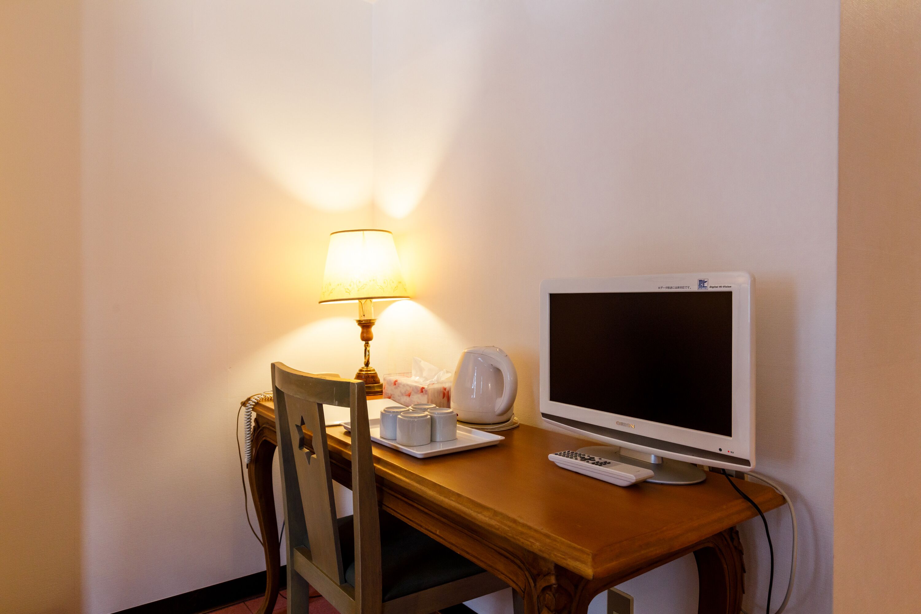 A hotel room desk with a computer monitor, a lamp, a chair, and a small table with a tissue box and a cup.
