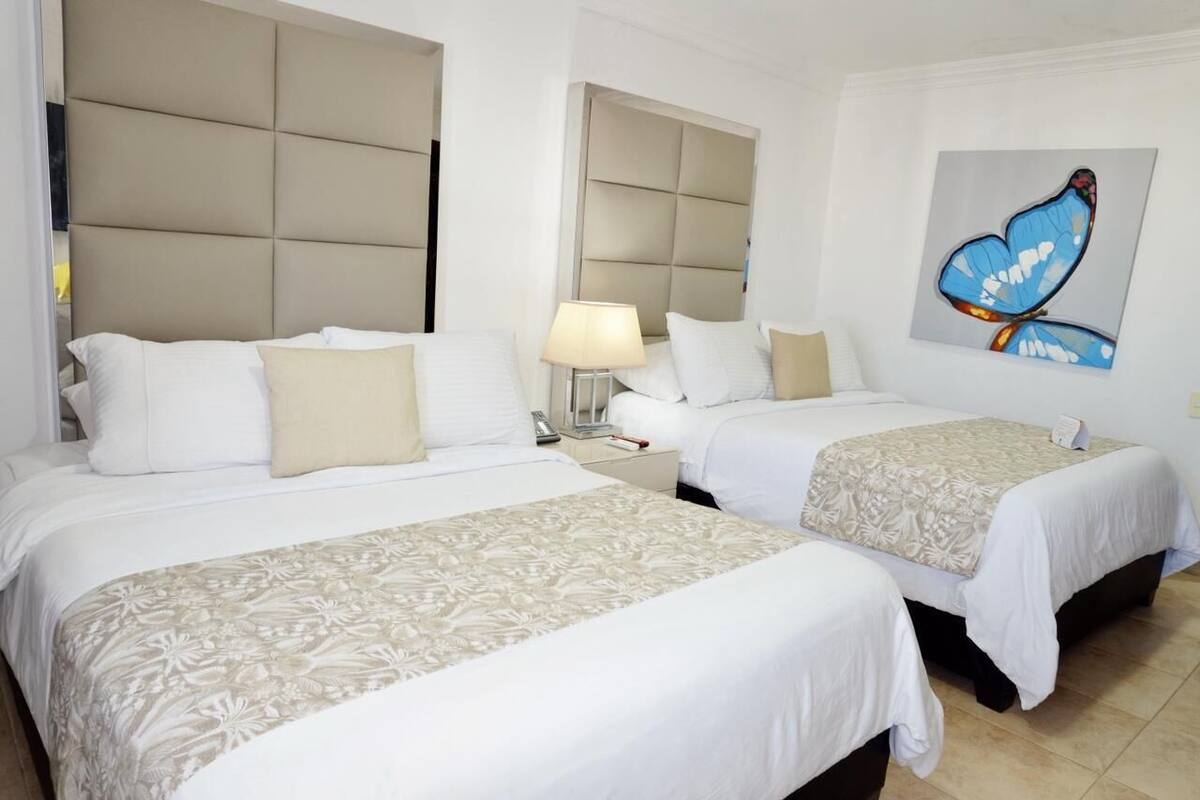 A hotel room with two beds, a large padded headboard, a bedside lamp, and a butterfly painting on the wall.