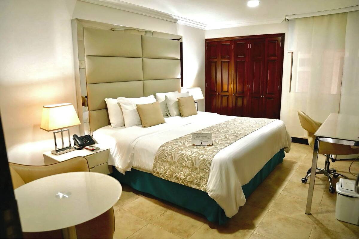 A hotel room with a large bed, a bedside table with a lamp, a chair, and a desk with a chair.