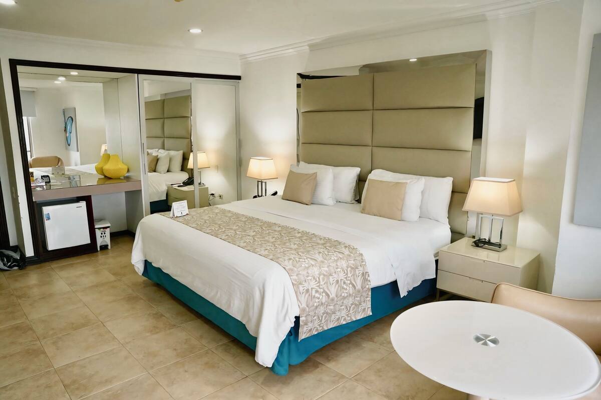 A hotel room with a large bed, bedside tables with lamps, a small desk with a lamp, a chair, and a round table.