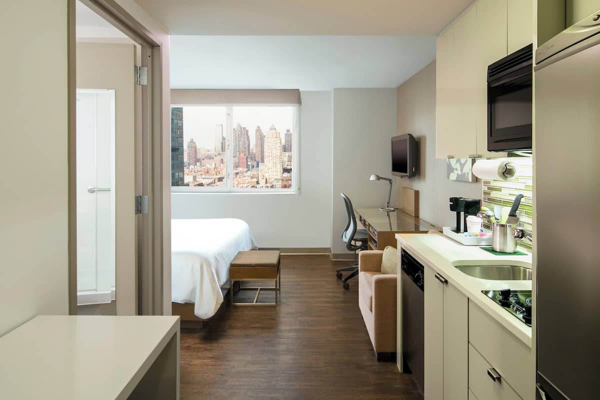 A compact hotel room with a bed, a small desk, a kitchenette, and a city view through a large window.