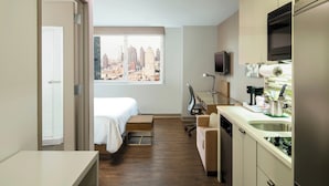 A compact hotel room with a bed, a small desk, a kitchenette, and a city view through a large window.