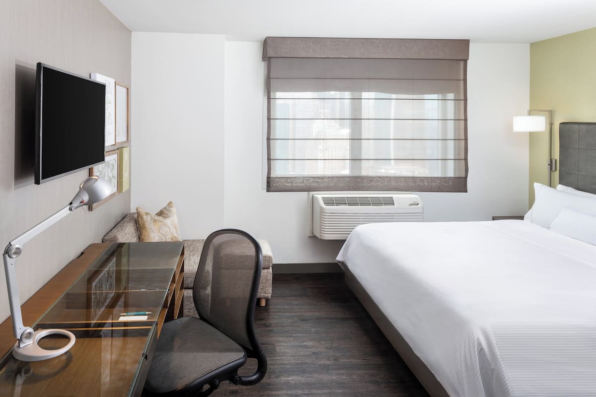 A hotel room with a bed, a desk with a chair, a TV, a window with blinds, and an air conditioning unit.