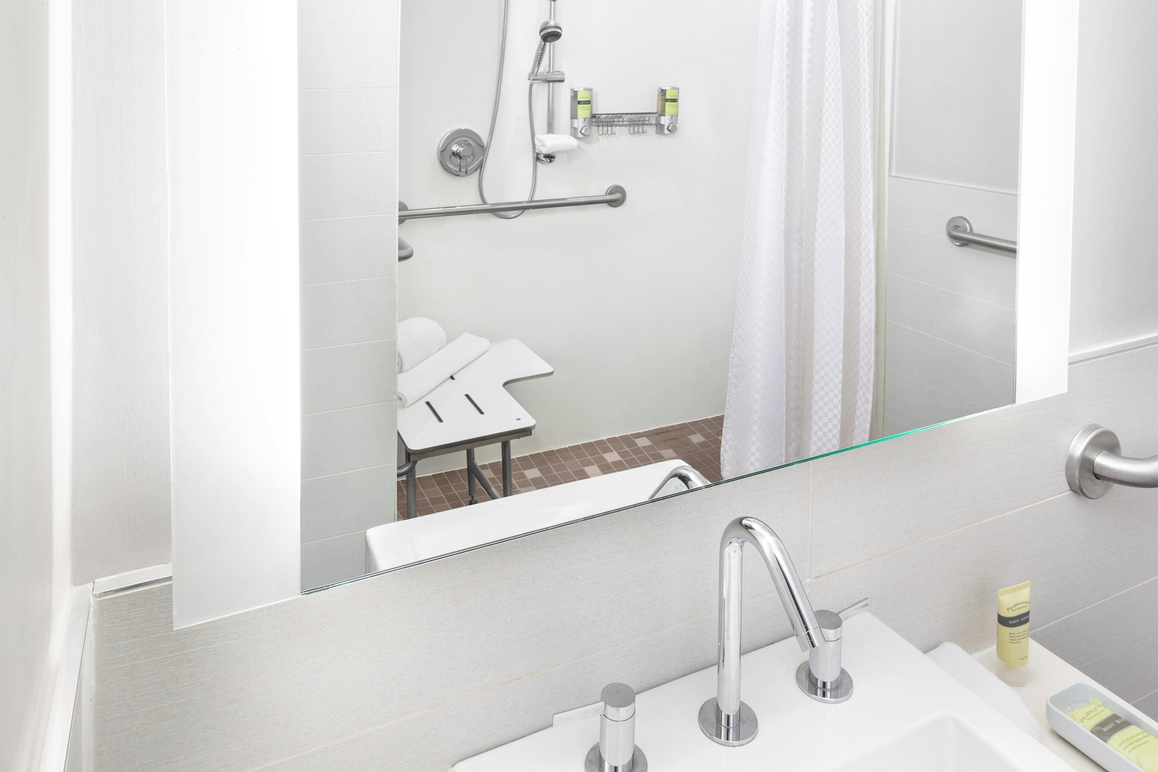Ein modernes Badezimmer mit einem großen Spiegel, einem Waschbecken mit zwei Wasserhähnen, einer Duschkabine mit Handbrause und einem WC mit Haltestange.