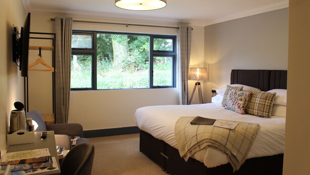 A hotel room with a bed, bedside table, lamp, and a view of greenery outside. - Woodlands Country House (Wigton)