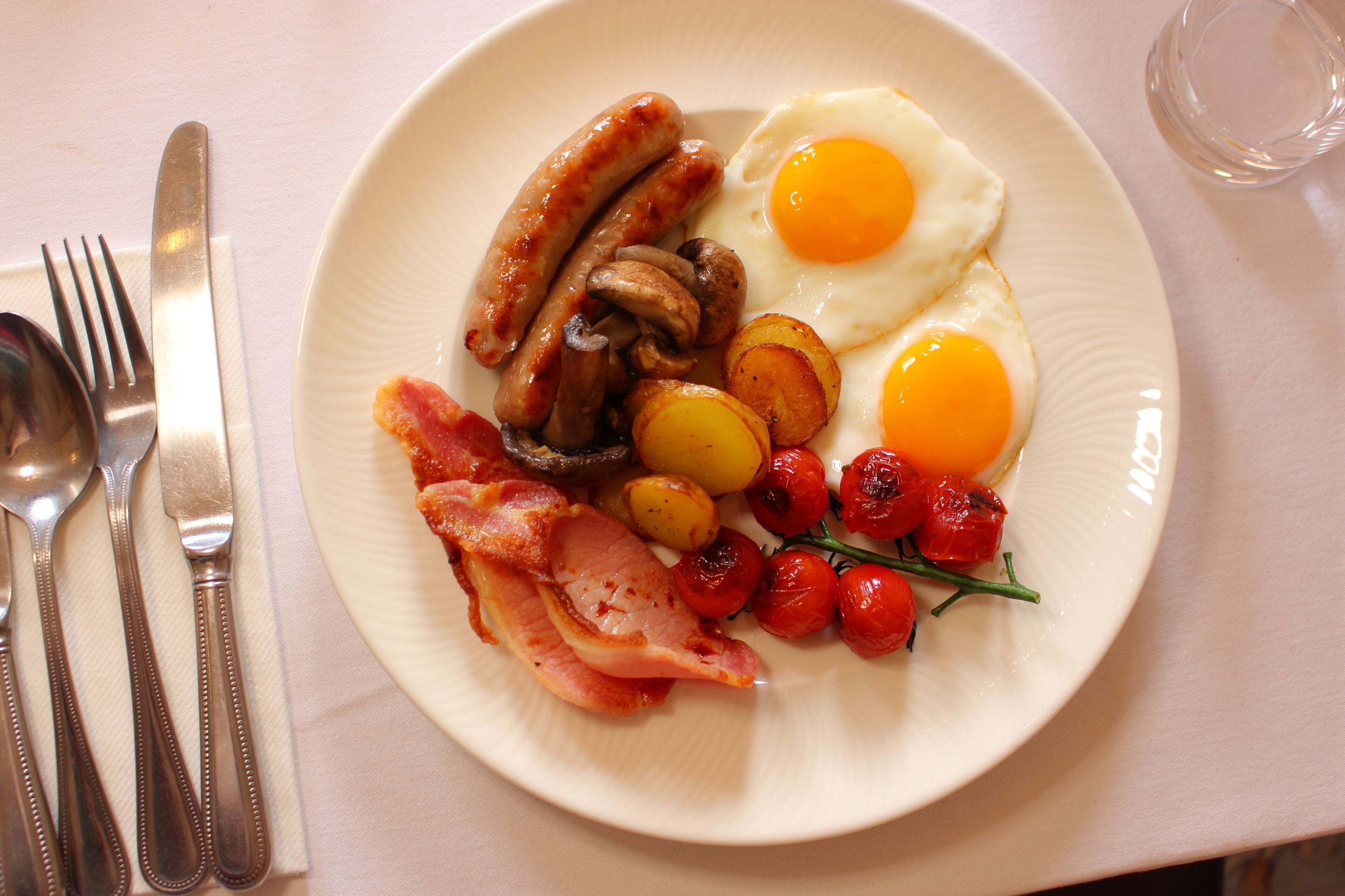 A plate of breakfast food including sausages, eggs, bacon, mushrooms, potatoes, and cherry tomatoes.