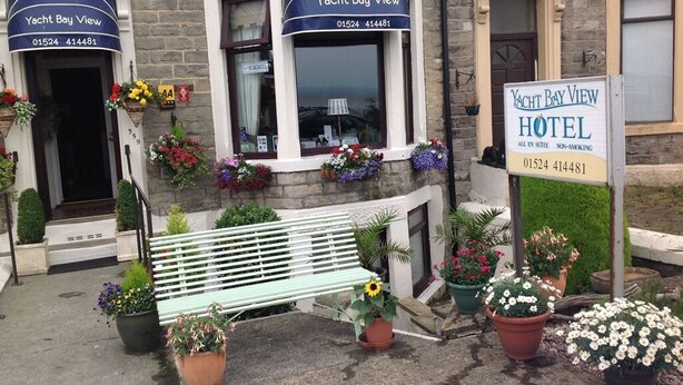 A building with a sign reading "Yacht Bay View Hotel" and a phone number. There is a bench and potted plants in front. - Yacht Bay View (Morecambe)