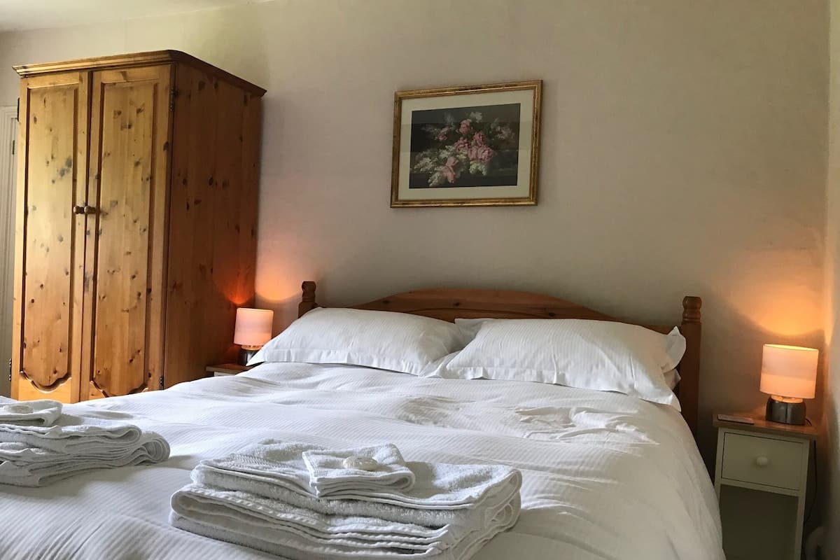 A neatly made bed with white linens and towels, a wooden wardrobe, and a framed floral painting on the wall.