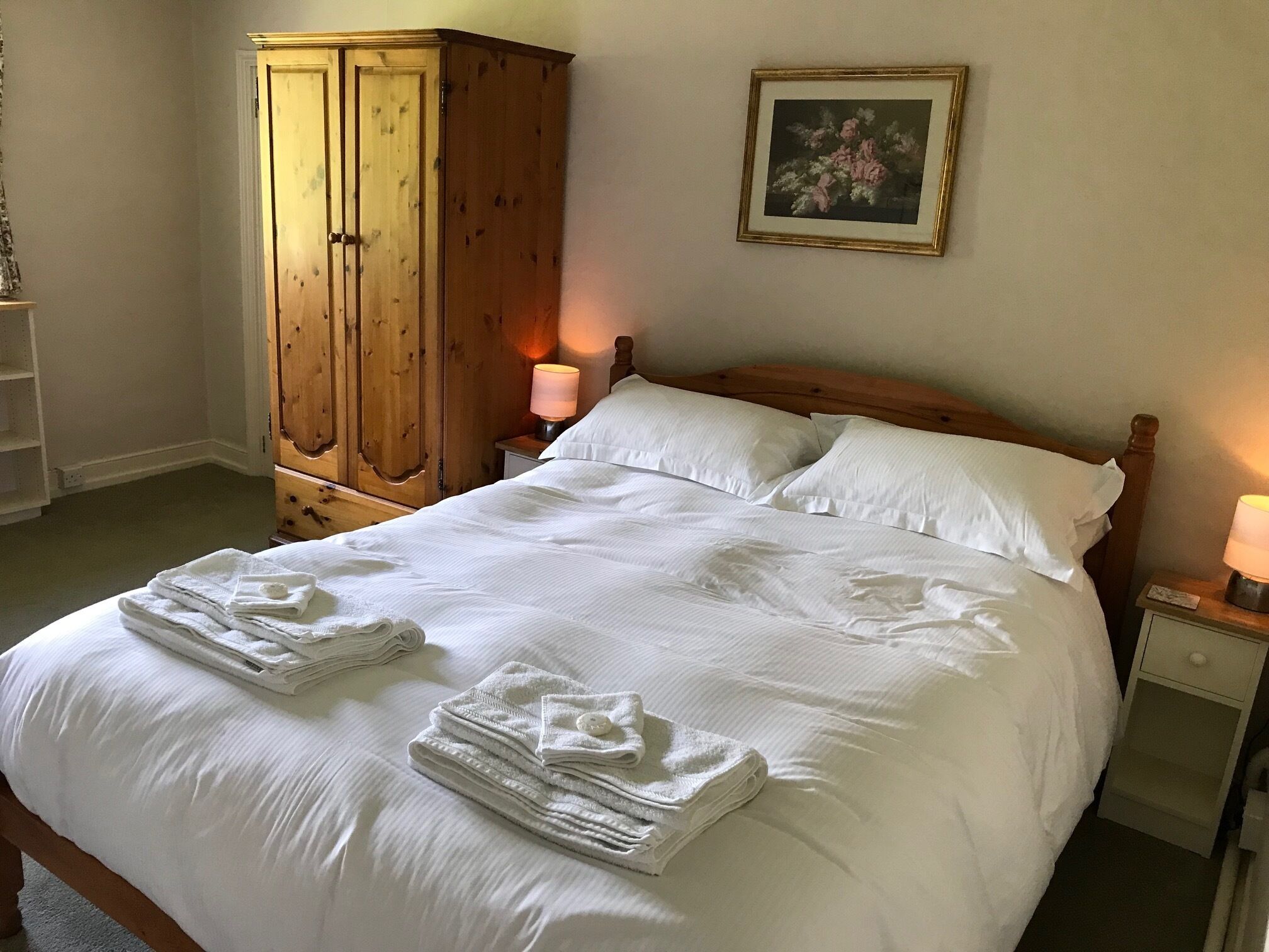 A neatly made bed with folded towels, a wooden wardrobe, a bedside lamp, and a framed picture on the wall.
