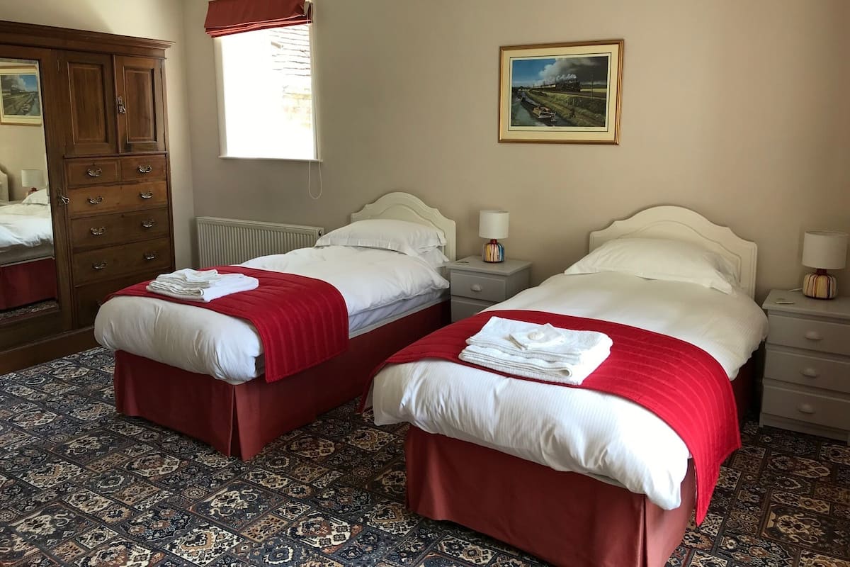 A hotel room with two beds, a large wooden wardrobe, a window with a view of the countryside, and a painting on the wall.