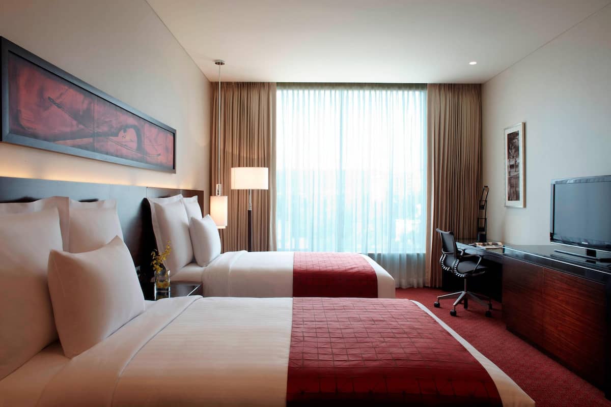 Habitación de hotel con dos camas, un escritorio, una silla, un televisor y un ventanal con cortinas.