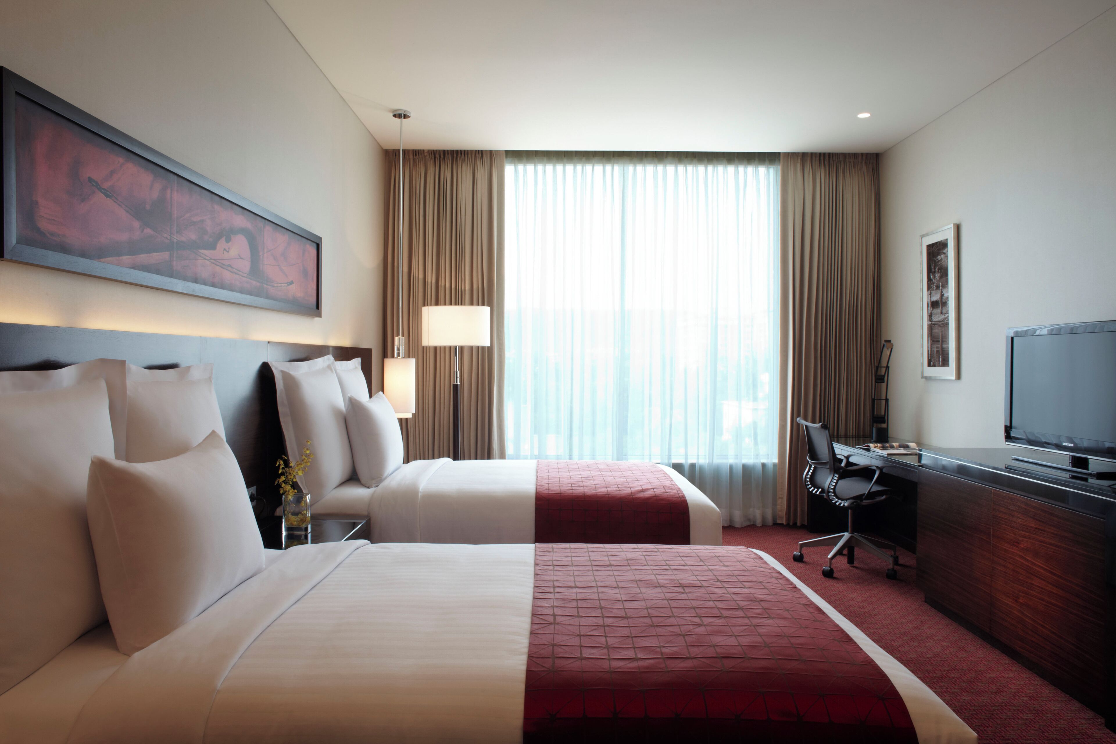 Una habitación de hotel con dos camas, un escritorio, un televisor y un ventanal grande con cortinas.