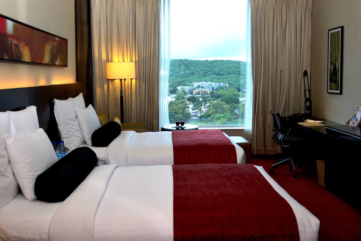 Habitación de hotel con dos camas, un escritorio, una silla, una lámpara y un ventanal con vistas a la ciudad.