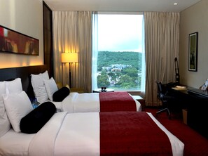 A hotel room with two beds, a desk, a chair, a lamp, and a large window offering a view of a cityscape. - JW Marriott Hotel Pune (Pune)