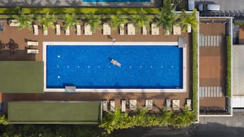 An aerial view of a hotel with a rectangular swimming pool surrounded by palm trees and lounge chairs.