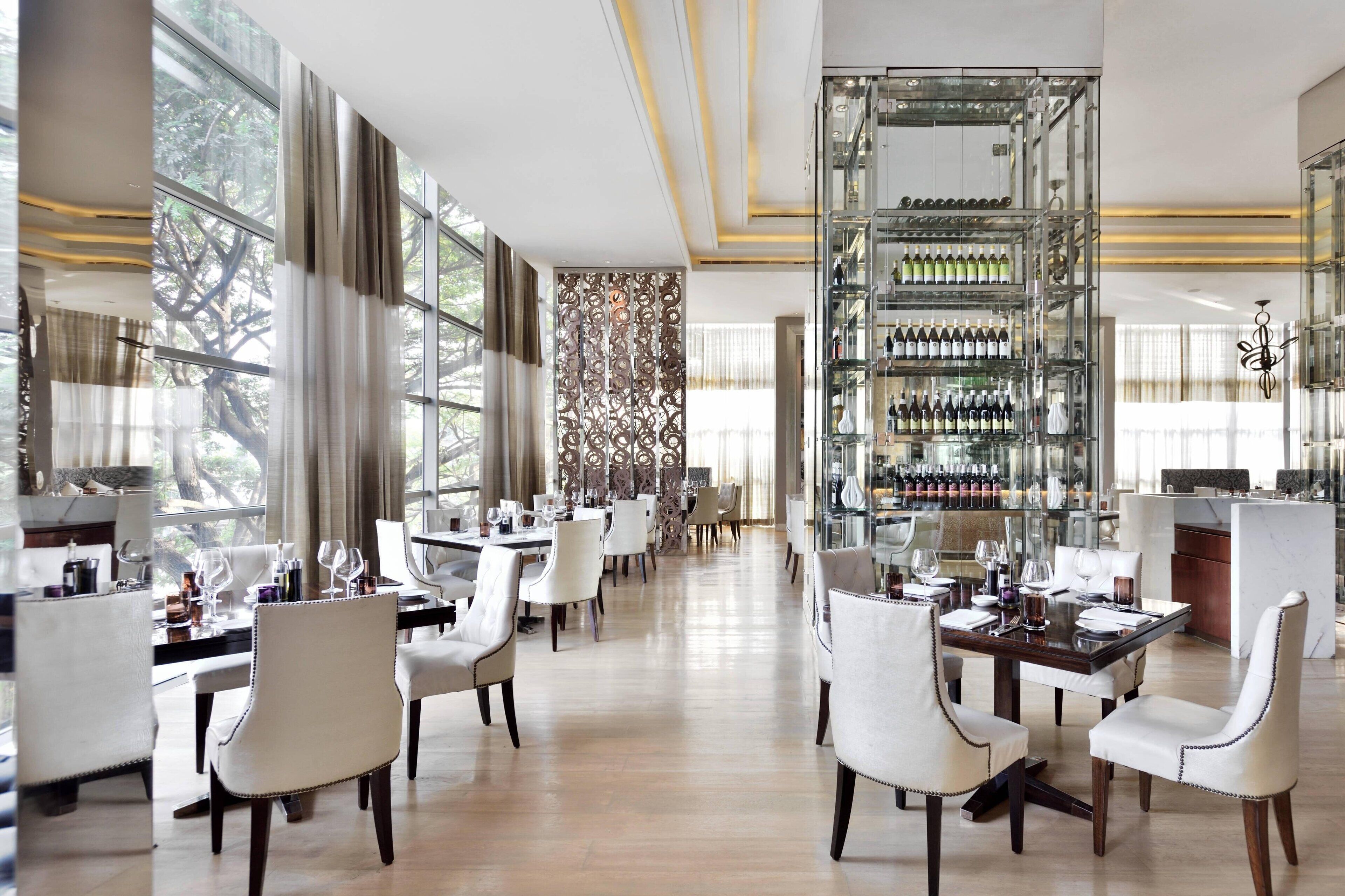 A spacious dining area with multiple tables and chairs, a wine rack with bottles, and large windows allowing natural light.