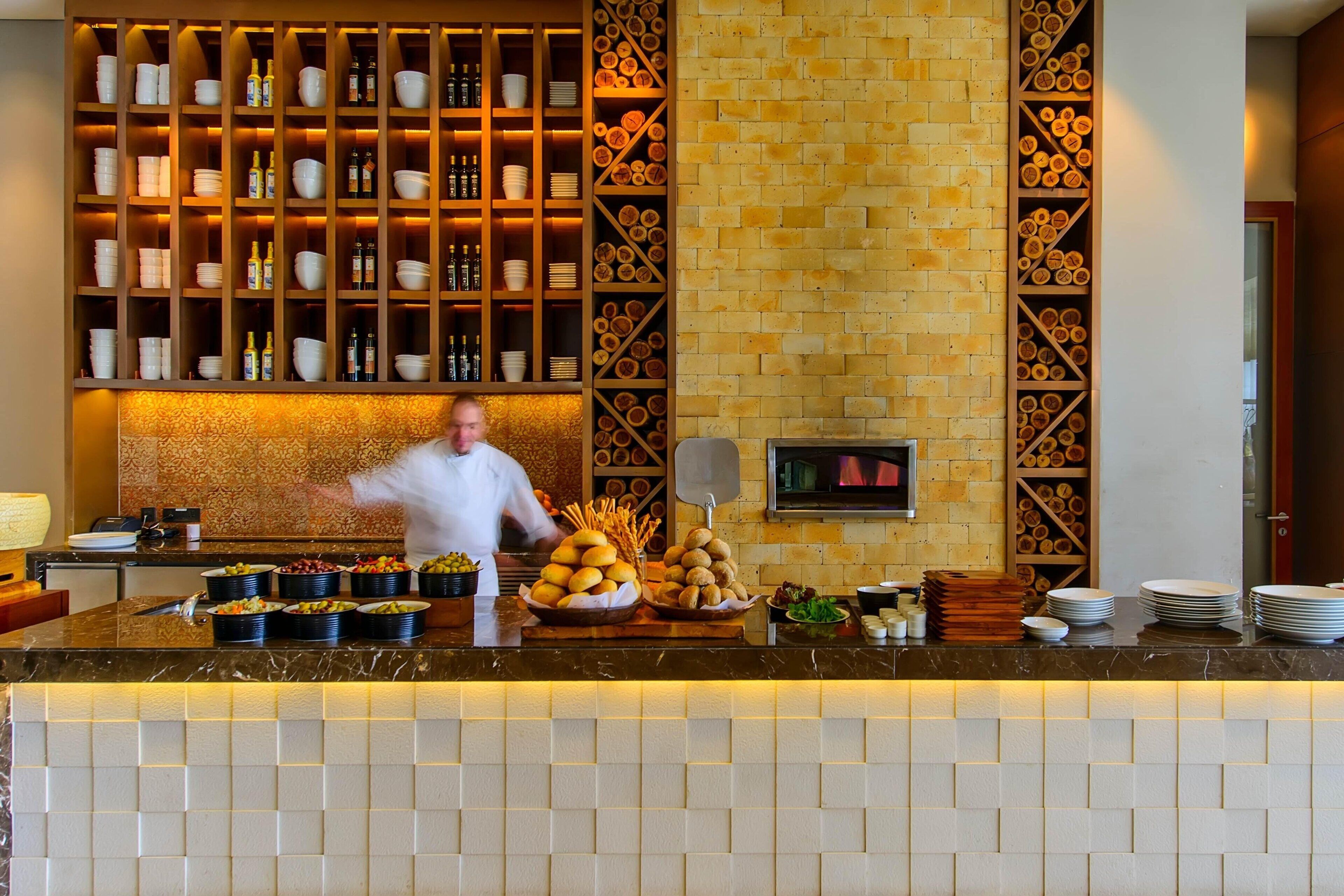 Un espace cuisine où un chef prépare des plats, un buffet proposant divers mets, et un mur de briques orné d’étagères remplies de bols et de bouteilles.