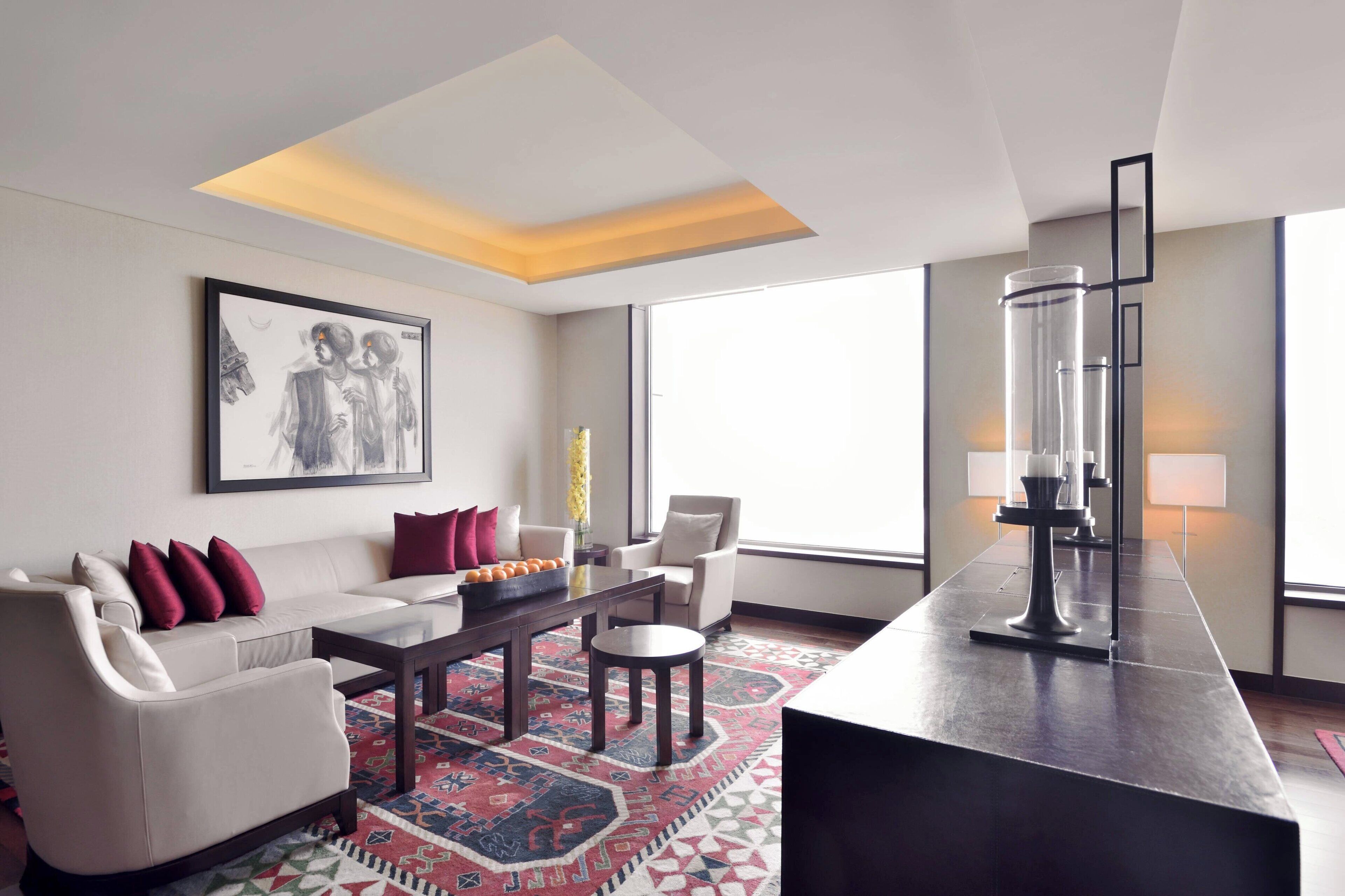 A modern living room with a sofa, armchairs, a coffee table, and a rug. There is a painting on the wall, a window with a view, and a tall vase on a table.