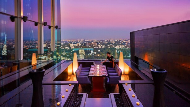 Un área de comedor en la azotea con una mesa para dos, rodeada de velas, y una vista panorámica de la ciudad de fondo.