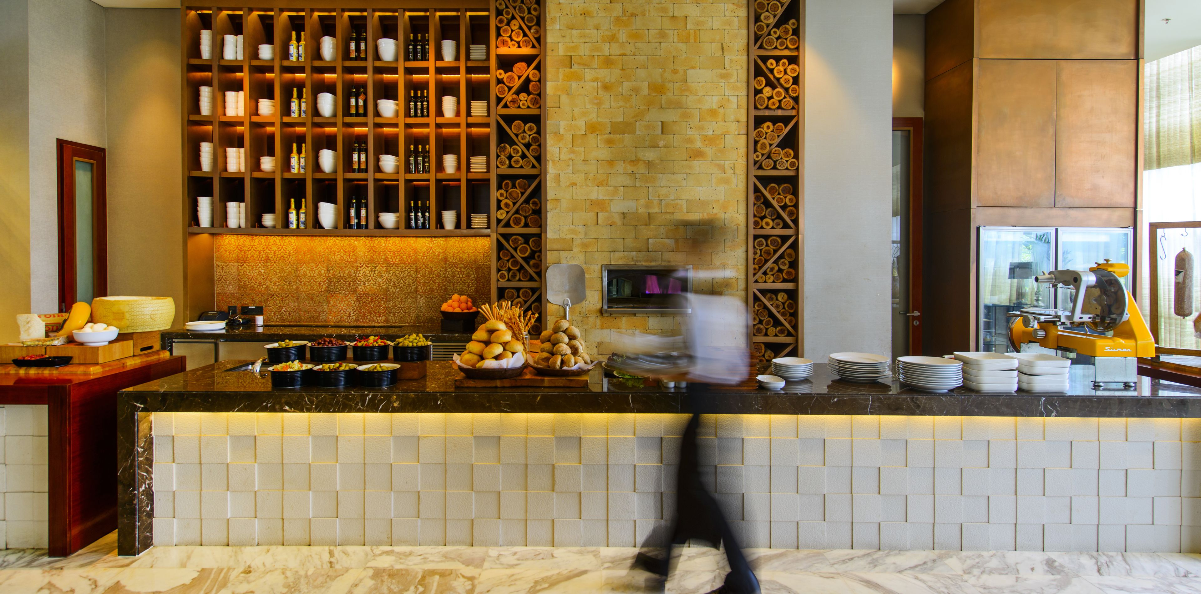 Un hall d’hôtel doté d’un comptoir de réception en marbre, d’une étagère murale avec des bols et des bouteilles, et d’un mur en briques sur lequel sont exposés des morceaux de bois.