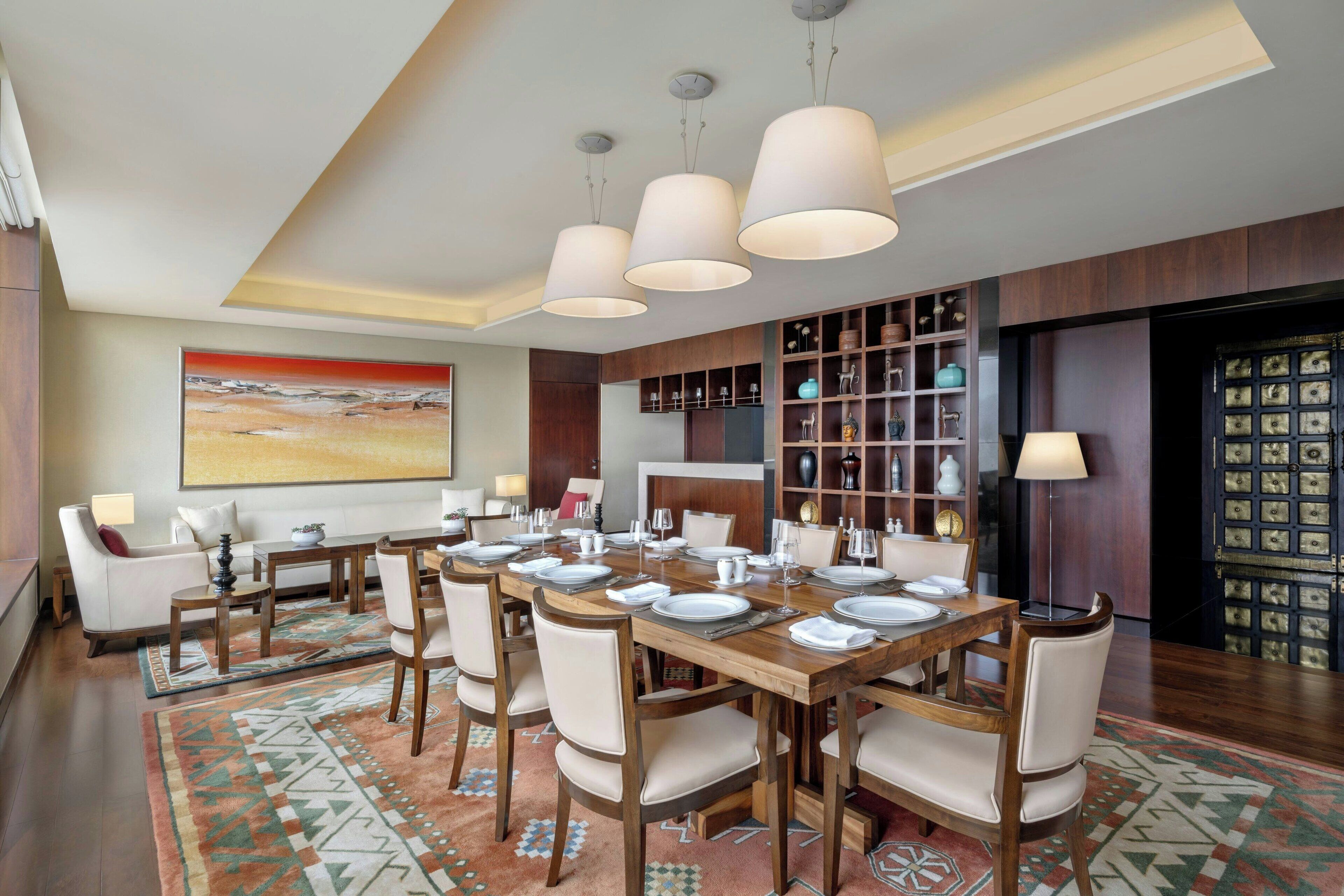 A dining area with a long wooden table set for six, surrounded by six chairs. There are hanging pendant lights above the table, and a large painting is visible on the wall to the left.