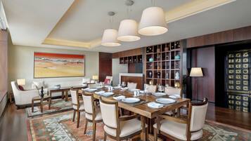 A dining area with a long wooden table set for six, surrounded by six chairs. There are hanging pendant lights above the table, and a large painting is visible on the wall to the left.