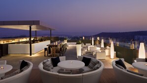 Un bar en la azotea con barra, zona de estar con mesas redondas y sillas acolchadas, y vistas a la ciudad al atardecer.