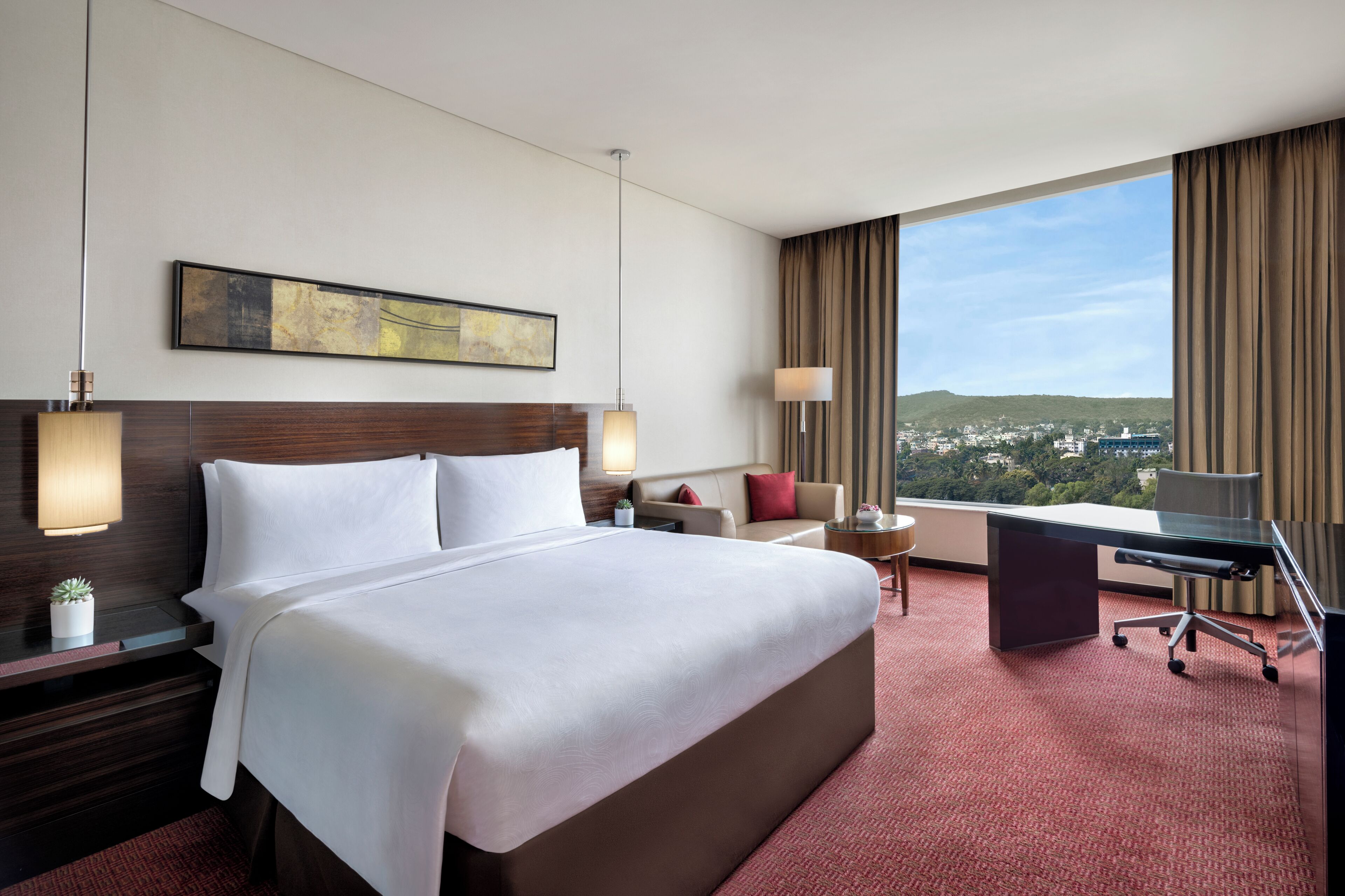 Habitación de hotel con una cama grande, un escritorio, una silla, una lámpara, una mesita con una planta y vistas a la ciudad.