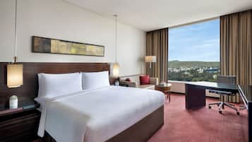 Habitación de hotel con una cama grande, un escritorio, una silla, una lámpara, una mesita con una planta y vistas a la ciudad.