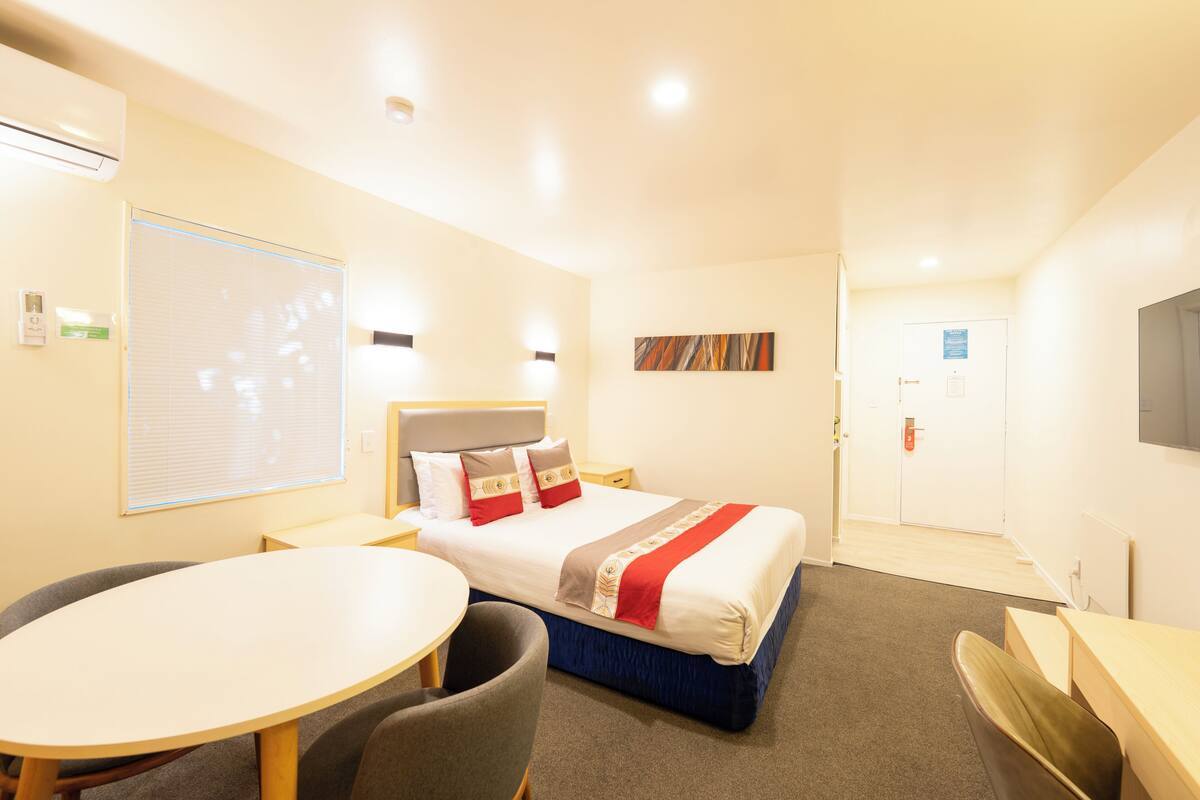 A hotel room with a bed, a desk, a chair, a small table, and a TV.