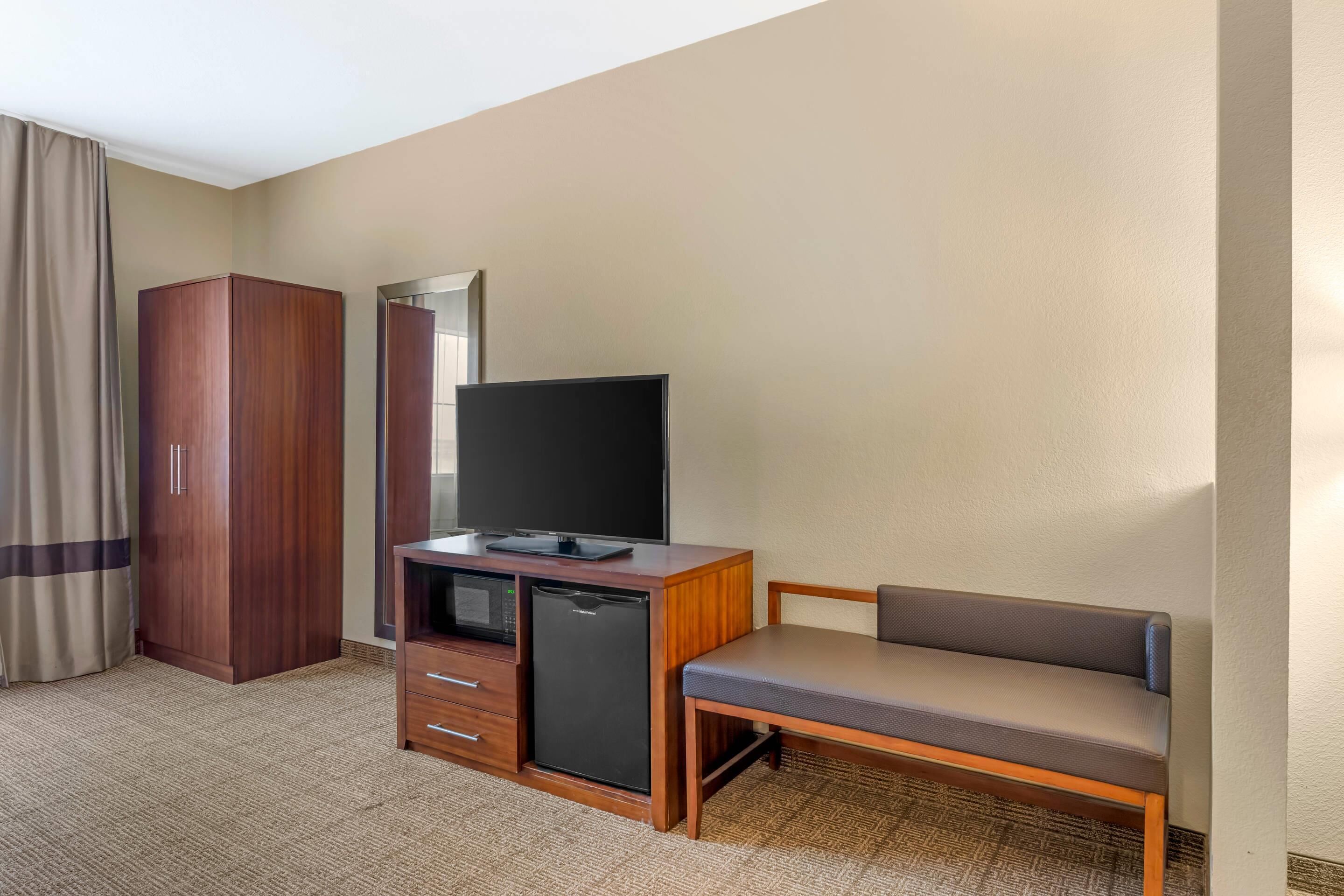 A hotel room with a wooden wardrobe, a small refrigerator, a flat-screen TV, and a bench.