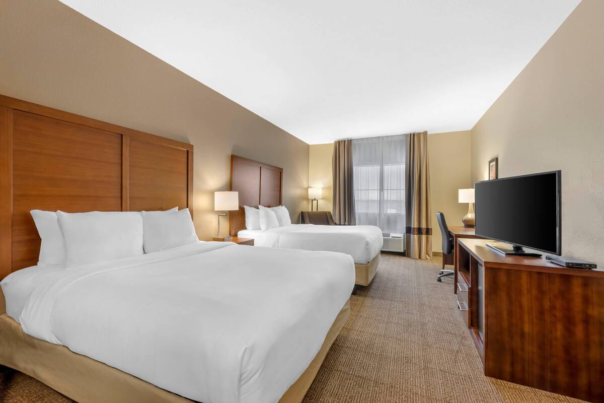 A hotel room with two beds, a desk, a television, and a large window with curtains.