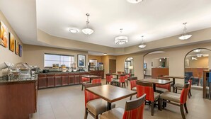 A hotel dining area with multiple tables and chairs, a kitchen area with a buffet, and a large window providing natural light. - Comfort Inn I-20 Midland Stanton (Stanton)