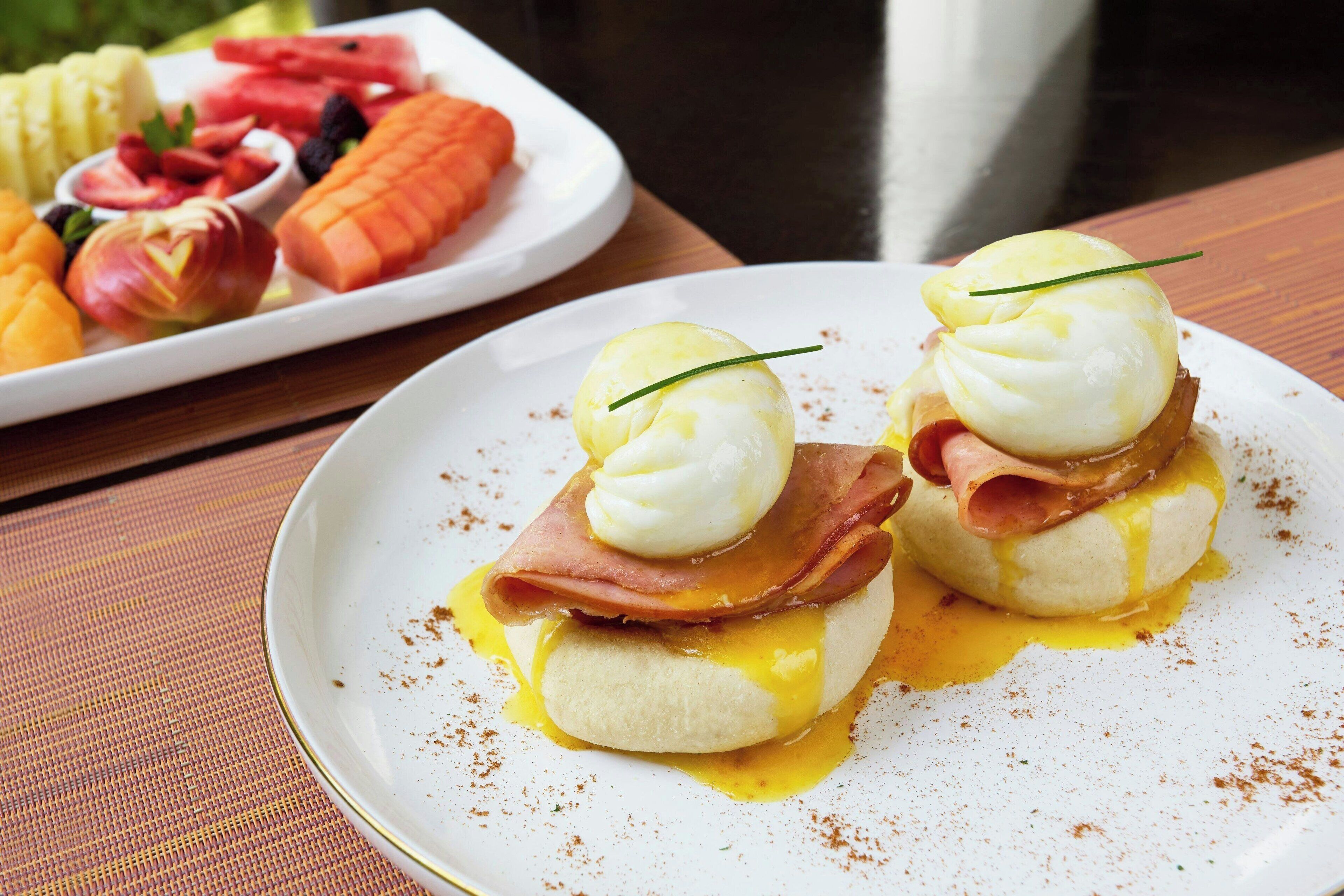 A breakfast plate with eggs Benedict and a side plate of fruit.