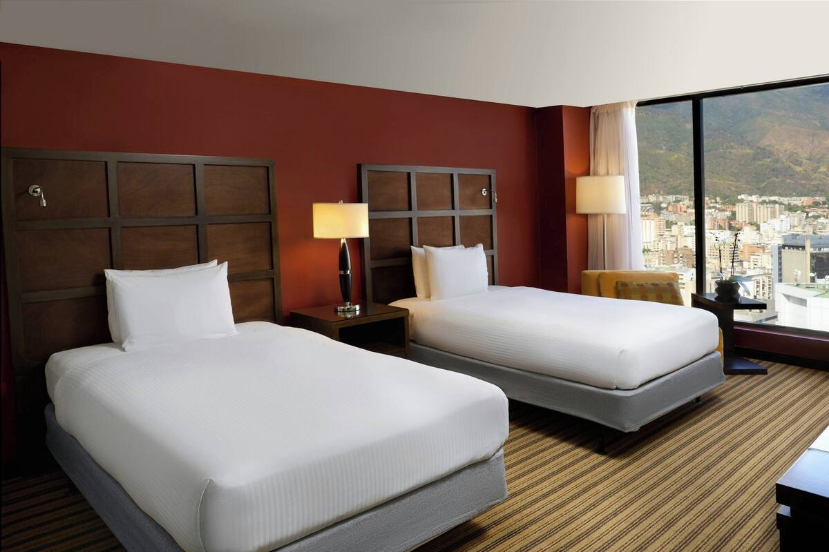 A hotel room with two beds, a large window with a city view, and a wooden headboard.
