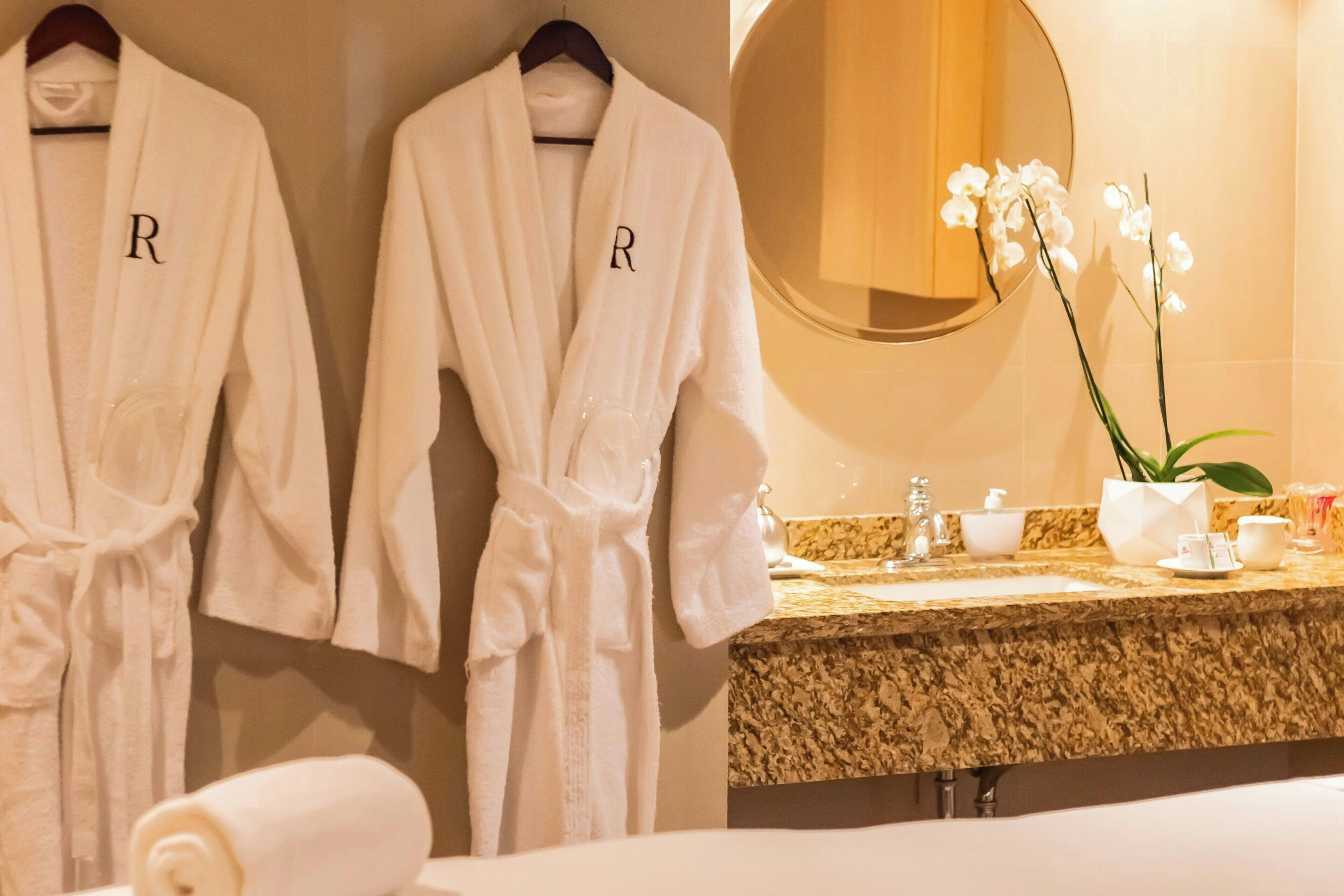 A hotel bathroom with two white robes hanging on a rack, a sink with toiletries, and a mirror.