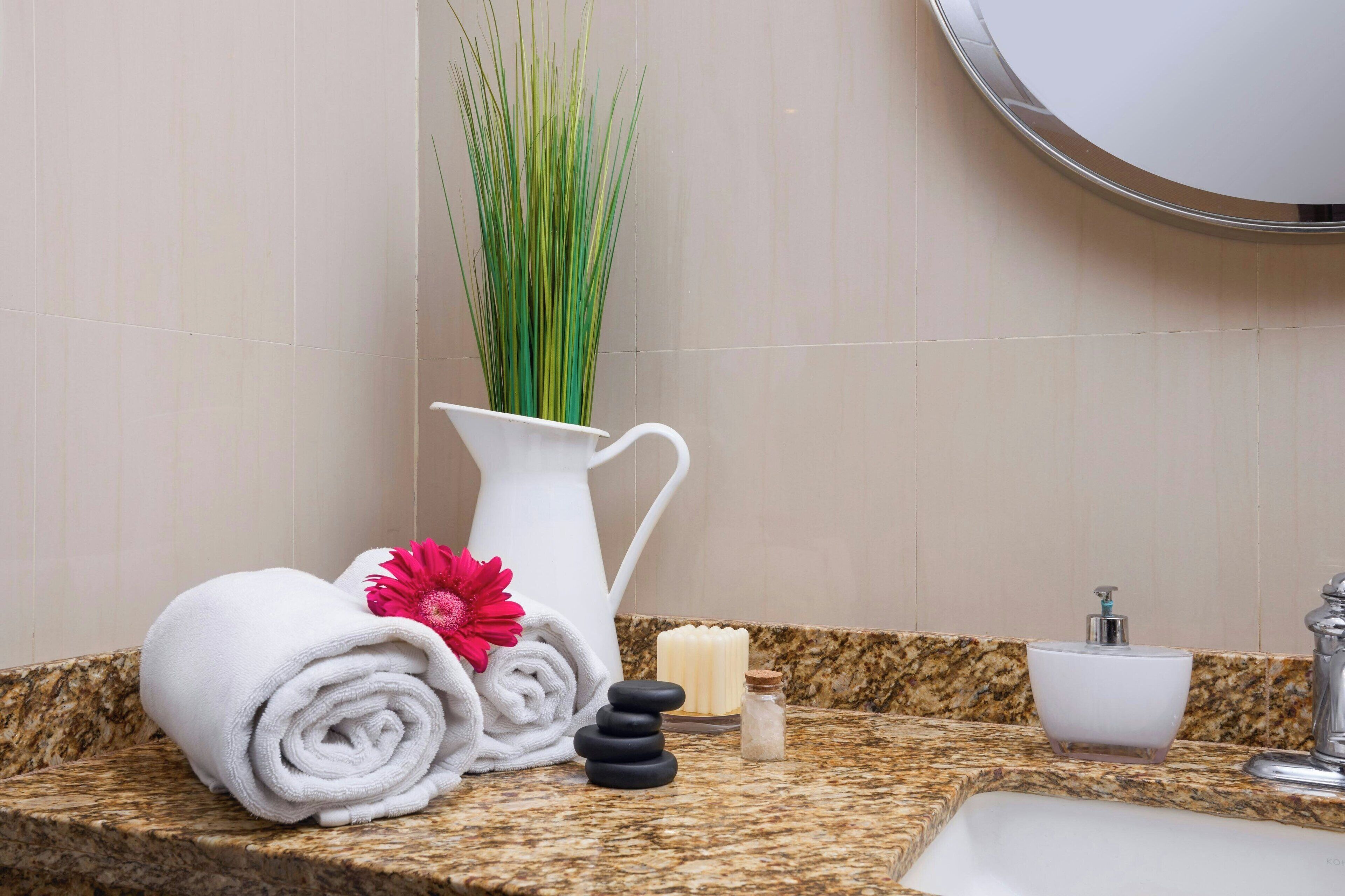 Une salle de bain avec un comptoir en marbre, un lavabo blanc, un miroir, un vase blanc contenant des plantes vertes, des serviettes enroulées, une fleur rose, un distributeur de savon et un petit flacon.