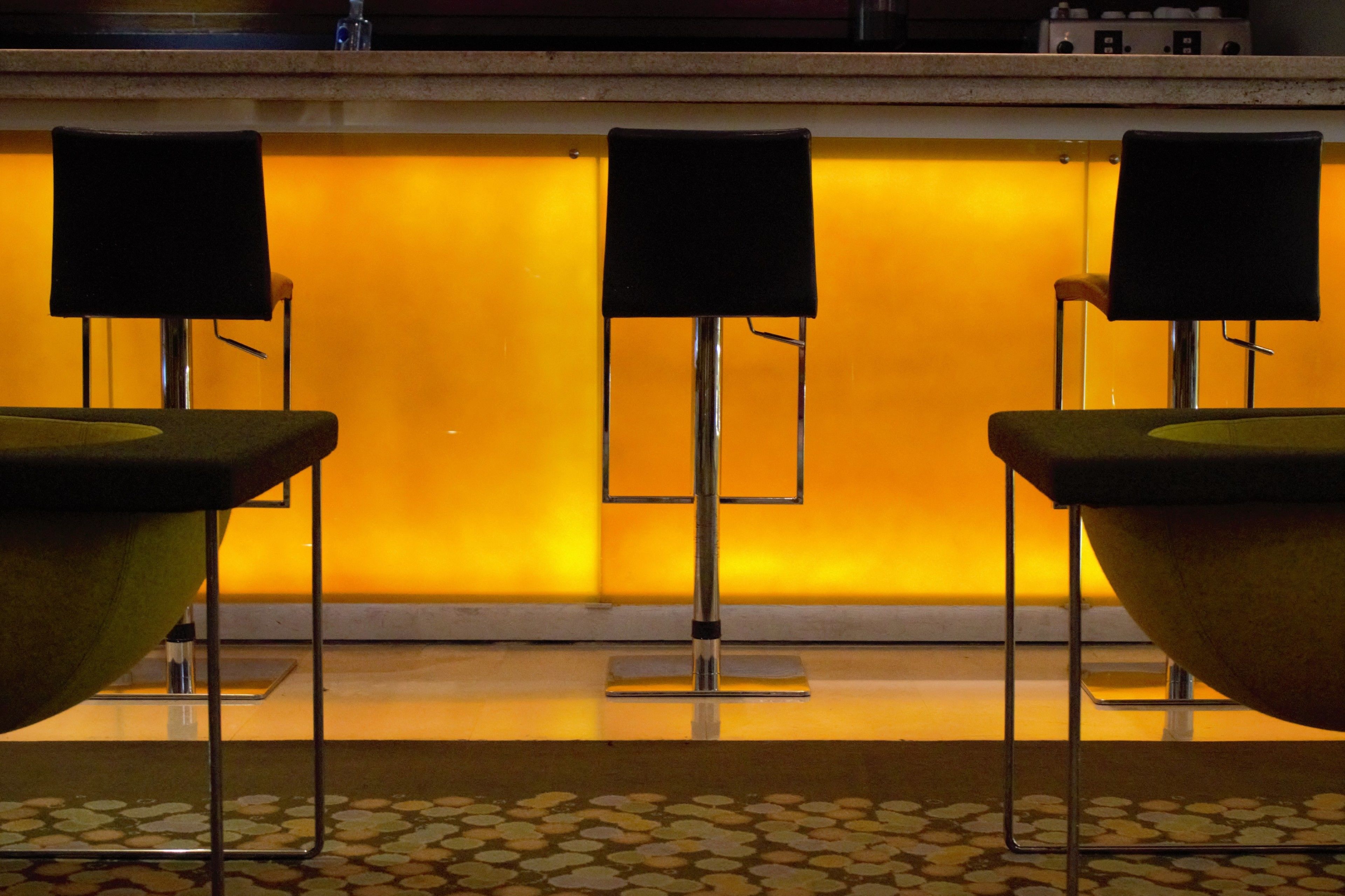 A bar area with three black bar stools and a yellow-lit wall.