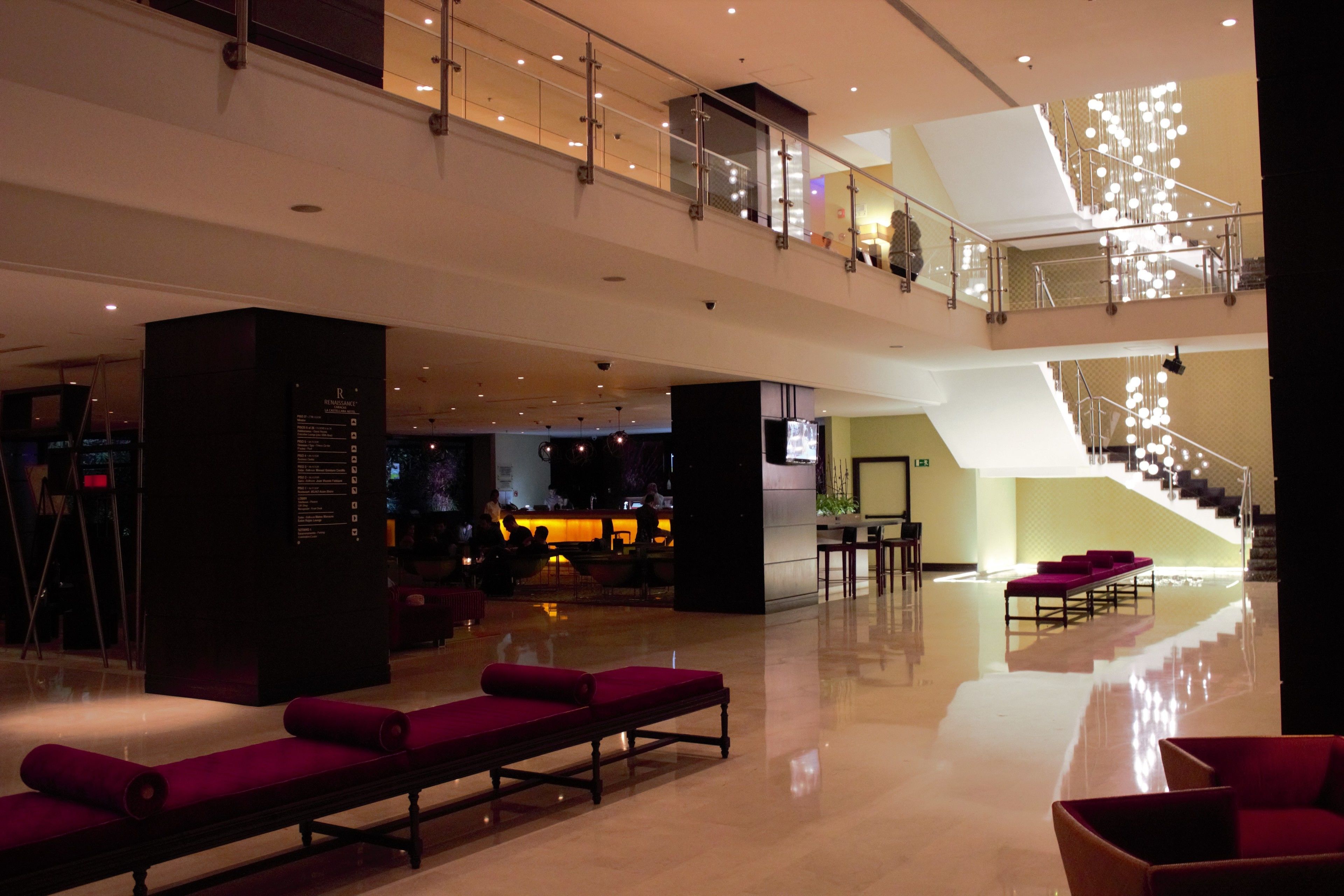 Un hall d’entrée spacieux au design moderne, comprenant un espace salon central avec des fauteuils rouges, un escalier avec des rampes en verre et un comptoir d’accueil.