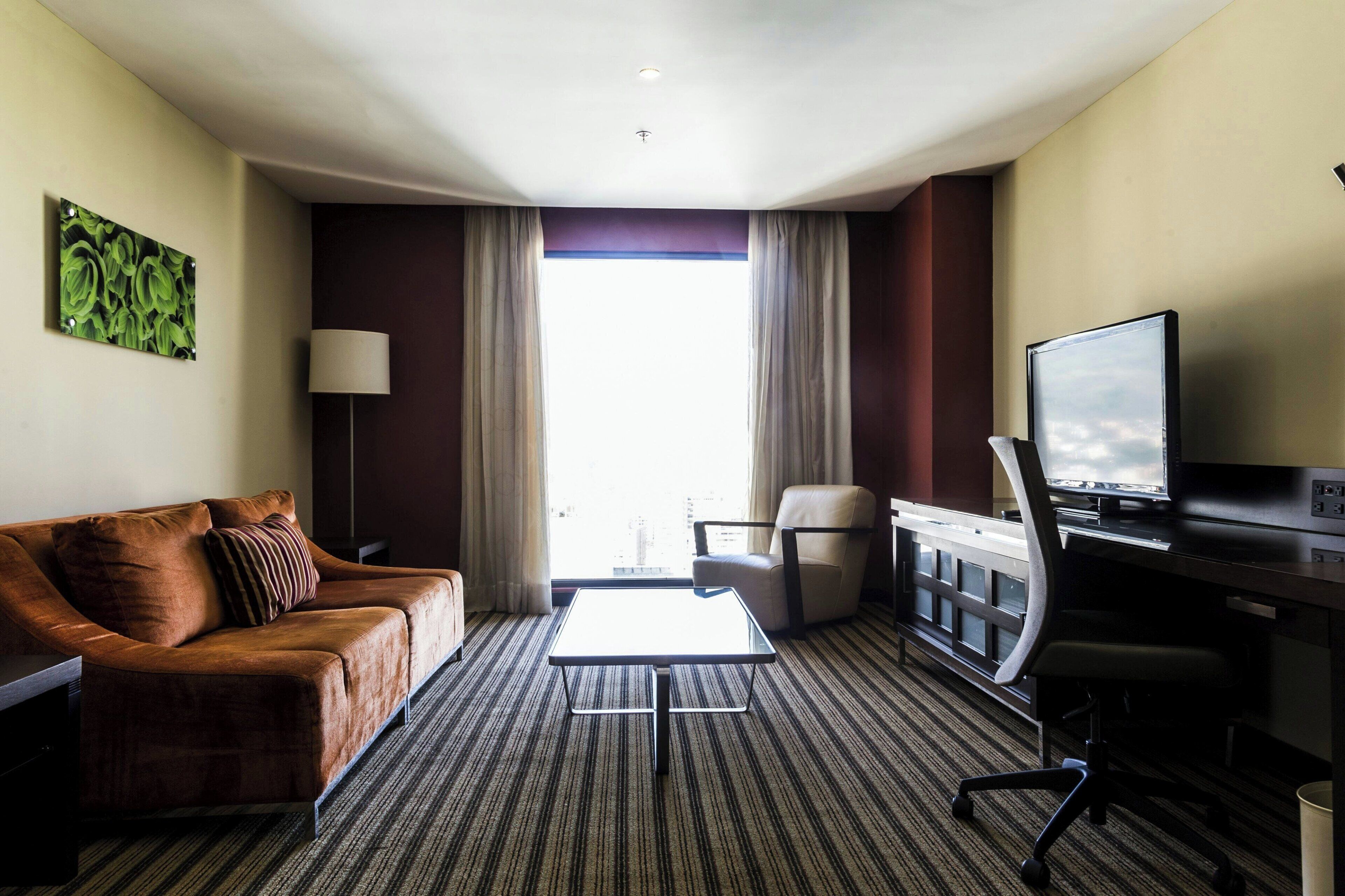 Ein Hotelzimmer mit Sofa, einem kleinen Tisch, einem Schreibtisch mit Computer, einem Sessel und einem Fenster mit Vorhängen.