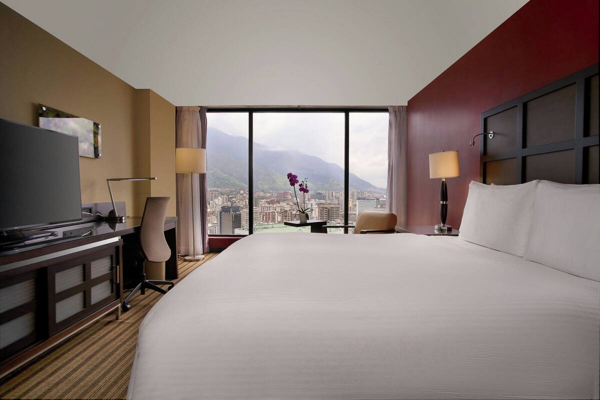 A hotel room with a large bed, a desk, a chair, a TV, and a view of a cityscape and mountains.