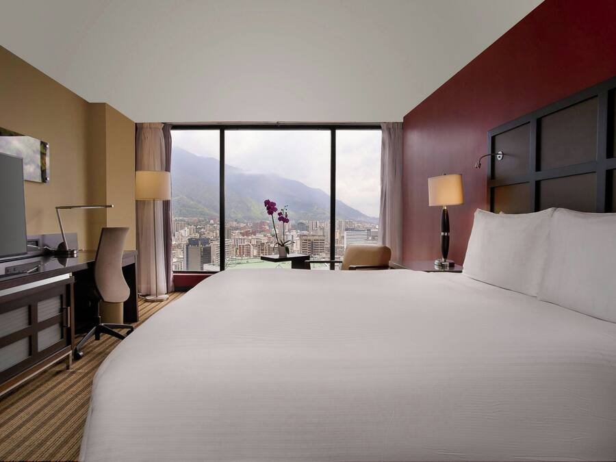 A hotel room with a large bed, a desk, a chair, a TV, and a view of a cityscape and mountains.