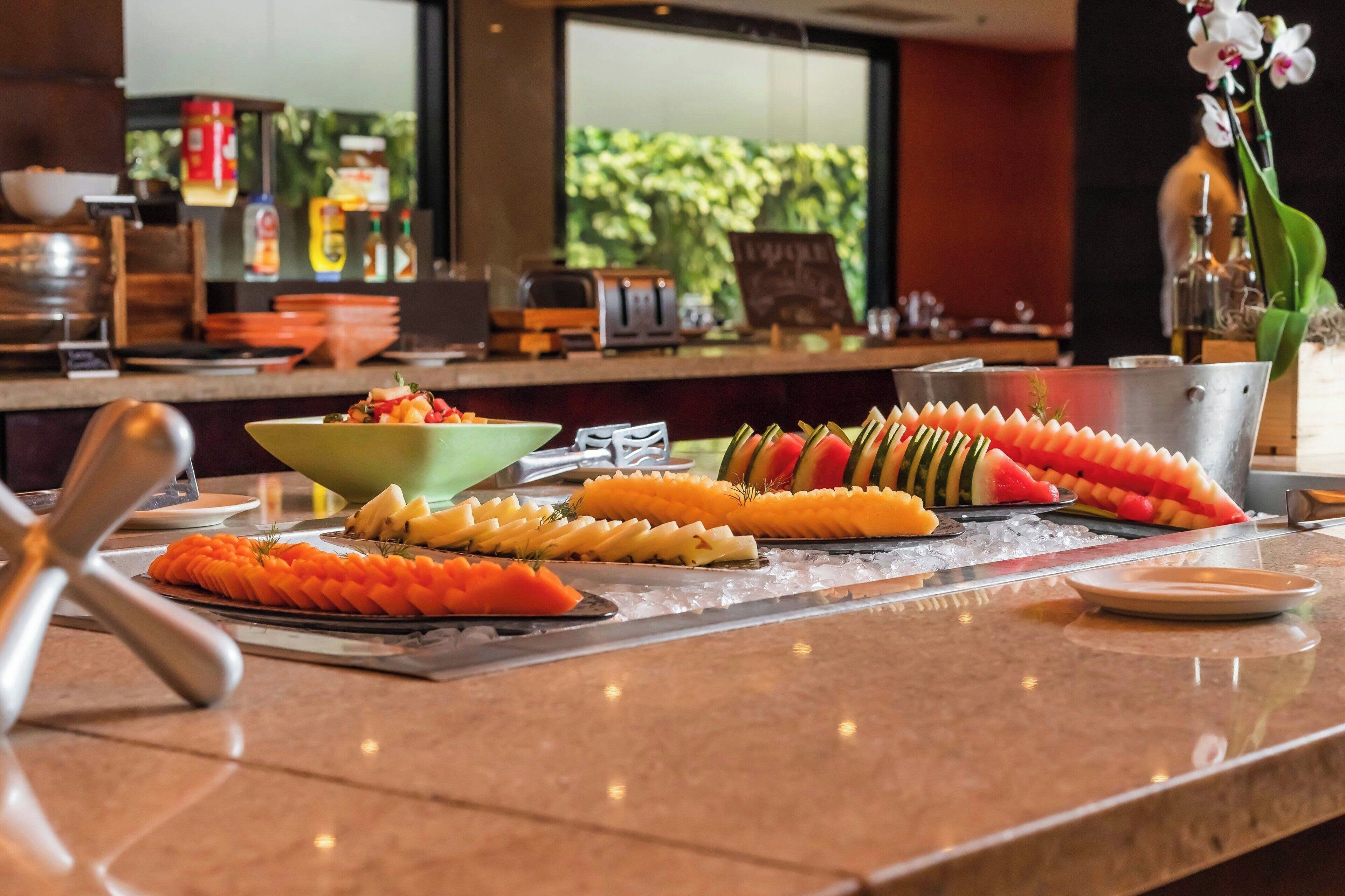 A buffet table with various fruits and vegetables, a coffee machine, and a view of greenery outside.