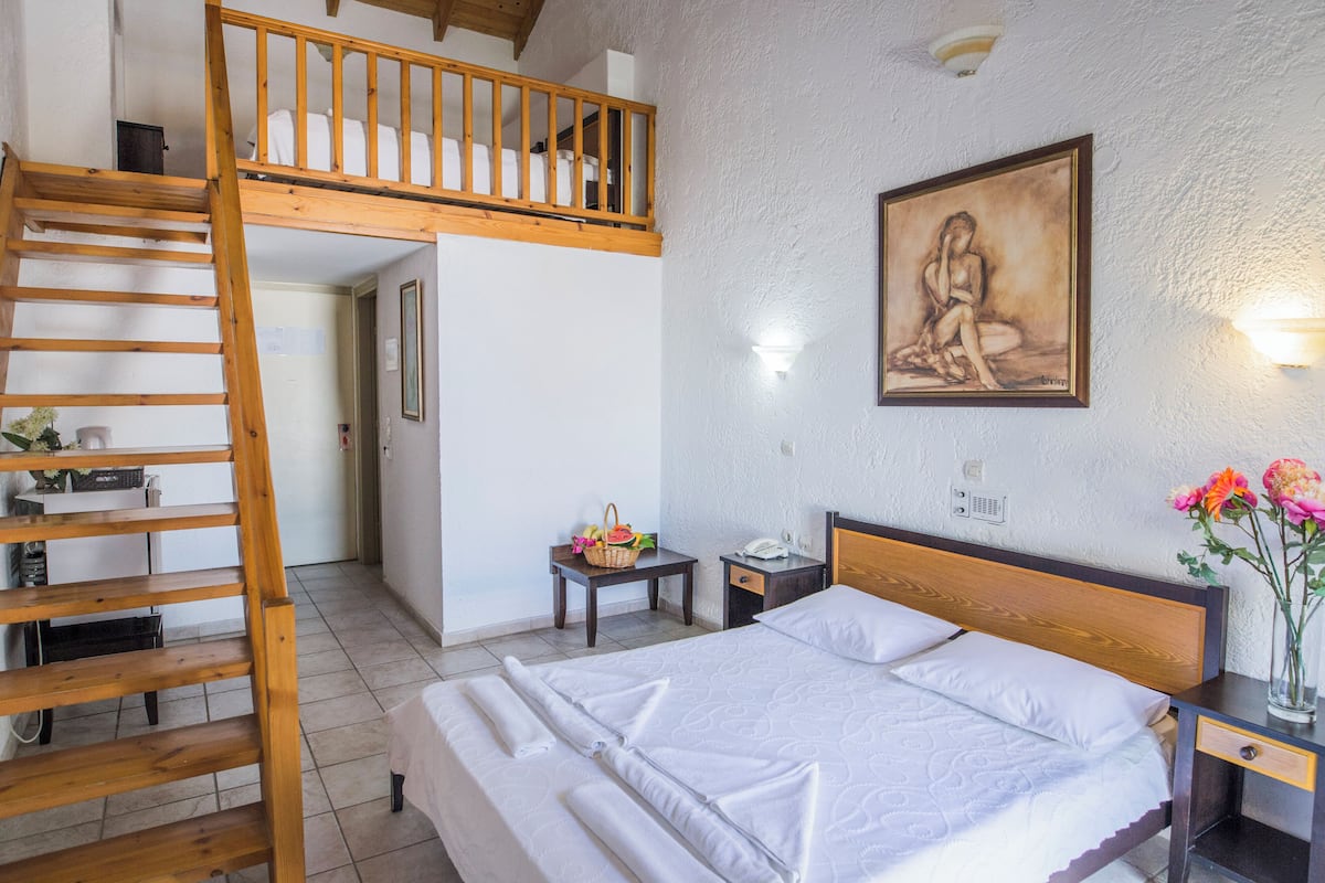 Een slaapkamer met een houten stapelbed, een enkel bed, een nachtkastje met een lamp, een schilderij aan de muur en een vaas met bloemen.
