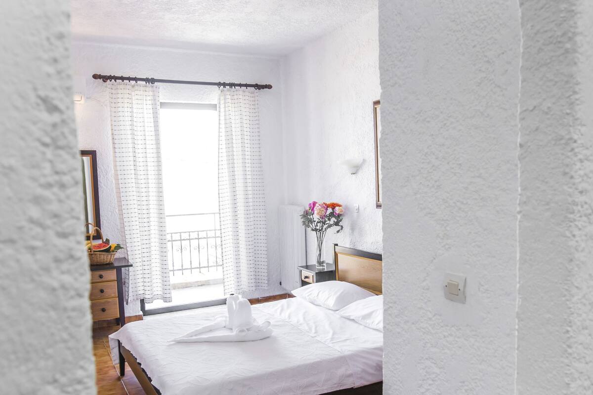 Een slaapkamer met een bed, een nachtkastje, een dressoir, een raam met gordijnen en uitzicht op het omliggende landschap.