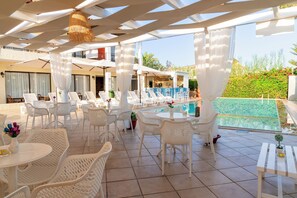 A poolside area with white wicker furniture, a bar, and a view of a pool. - Creta Aquamarine hotel (Rethymno)