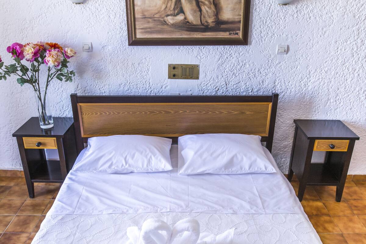 Een net opgemaakt bed met witte lakens, een houten hoofdbord en een schilderij erboven. Er staan twee nachtkastjes met lampen en op één nachtkastje staat een bloemenvaas.