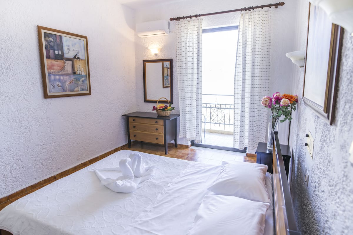 Een slaapkamer met een bed, een dressoir, een spiegel, een schilderij en een raam met gordijnen.