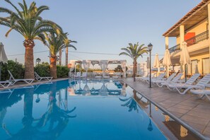 A swimming pool with white lounge chairs and palm trees, surrounded by a building with balconies and outdoor seating. - Creta Aquamarine hotel (Rethymno)