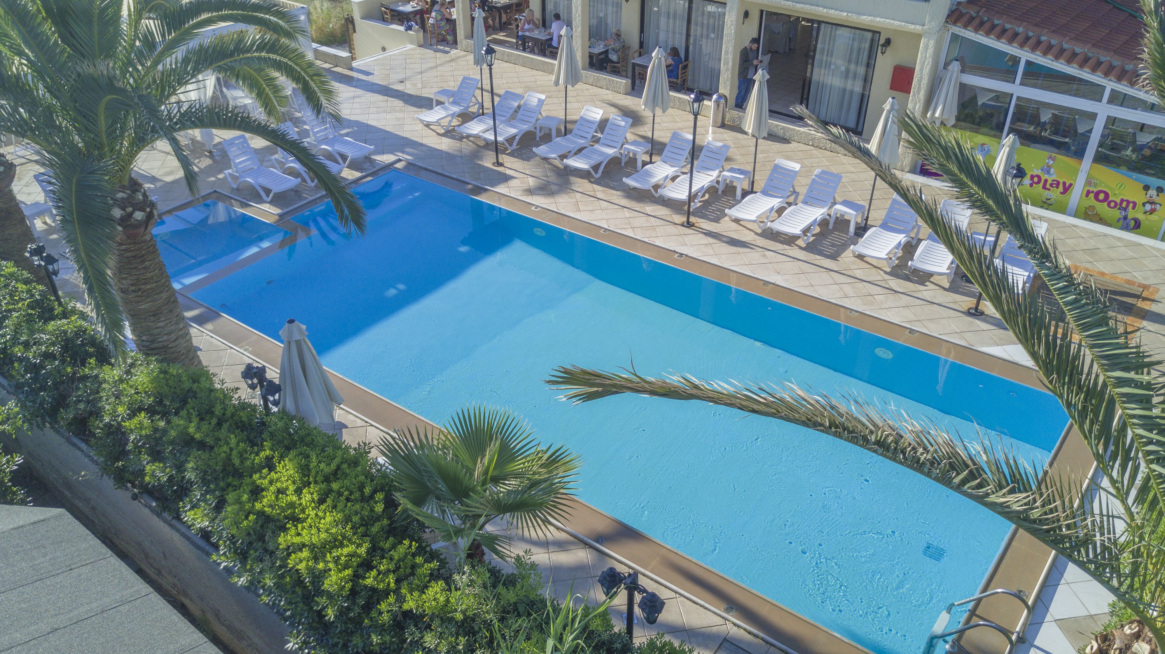 A swimming pool with lounge chairs and umbrellas, surrounded by palm trees and a building with a Play Room sign.
